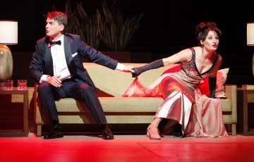 Smokkiin pukeutunut mies istuu sohvalla vasemmalle katsoen ja pitää kädestä kiinni lumoavaan pukuun pukeutunutta naista, joka nojaa poispäin ja näyttää yllättyneeltä; molemmat näyttävät olevan teatterin lavalla.