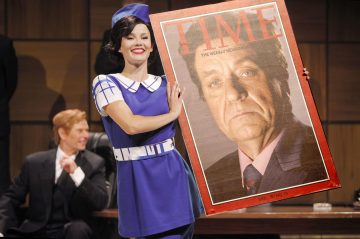 Siniseen retropukuun pukeutunut nainen hymyilee ja pitää kädessään suurta rekvisiittaa TIME-lehden kannesta, jossa on vakava, pukuinen vanhempi mies. Taustalla istuu pukuinen mies hymyillen. Kohtaus näyttää olevan näytelmästä.