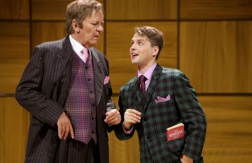 Lavalla esiintyy kaksi värikkäisiin pukuihin pukeutunutta miestä; vanhempi mies raidallisessa puvussa näyttää tyytymättömältä, kun taas nuorempi mies ruudullisessa puvussa hymyilee ja pitää kädessään punaista kirjaa, jossa lukee RAHA. Taustalla on puupaneeleita.