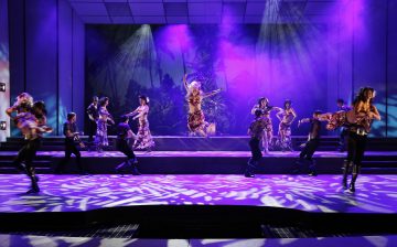 Esiintyjät tanssivat energisesti kirkkaasti valaistulla lavalla, jossa on violetteja ja sinisiä valonheittimiä. Yksi tanssija hyppää keskellä ilmaa, kun taas muut poseeraavat dynaamisesti heidän ympärillään luoden eloisan, vilkkaan kohtauksen.