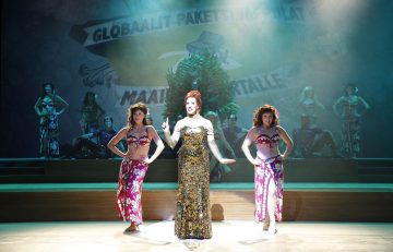 Keskellä näyttämöä seisoo kimaltelevaan pukuun pukeutunut esiintyjä, jota reunustaa kaksi kukka-asuihin pukeutunutta tanssijaa. Heidän takanaan istuu muita tanssijoita, ja taustalla on dramaattinen valaistus ja suuri banderolli.