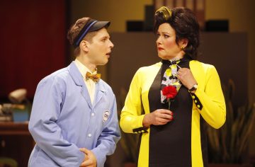 Siniseen univormuun ja rusettisolmioon pukeutunut mies puhuu naiselle, joka on pukeutunut kirkkaan keltaiseen ja mustaan asuun, jossa on kukkahuivi. Nainen näyttää yllättyneeltä ja pitää kättään rintaansa vasten. Molemmat ovat näyttämöllä teatterikohtauksessa.