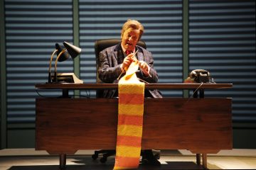 Henkilö istuu suuren puisen työpöydän ääressä, kädessään pitkä, värikäs neulottu huivi. Työpöydällä on lamppu, puhelin ja papereita. Vaakasuorat kaihtimet peittävät takana olevan ikkunan, ja henkilö näyttää keskittyneen neulomiseensa.