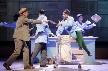 Ryhmä postityöntekijöiksi pukeutuneita näyttelijöitä lavalla käsittelee kirjeitä ja paketteja. Yksi sinipukuinen henkilö istuu kärryllä, kun taas toinen pukuun pukeutunut henkilö ojentaa kirjekuoren. Taustalla on neliönmuotoisista paneeleista koostuva seinä.