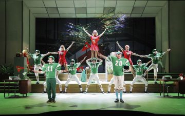 Jalkapalloilijoiksi ja cheerleadereiksi pukeutunut teatteriryhmä poseeraa energisesti lavalla, ilmassa on konfettia ja vihreät peliasut ja viirit. Lavastuksessa on modernit kalusteet ja kirkas, dramaattinen valaistus.