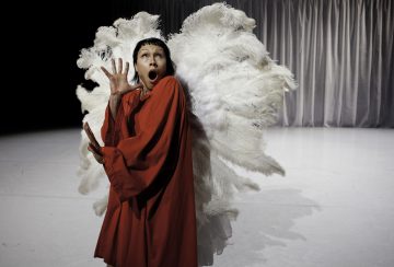 Kirkkaanpunaiseen asuun pukeutunut henkilö poseeraa dramaattisesti yllättynyt ilme kasvoillaan suurten valkoisten höyhensiipien edessä. Tausta on minimaalinen vaaleilla verhoilla.