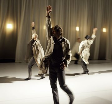 Neljä trenssitakkiin pukeutunutta ihmistä esiintyy kirkkaasti valaistulla näyttämöllä, jossa on beigen sävyiset verhot ja seinävalot, ja kukin heistä nostaa yhden käden liikkeessä, mikä luo dynaamisen liikkeen ja teatterillisen ilmaisun tunteen.