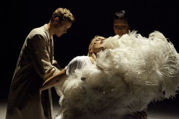 Lavalla esiintyy kolme ihmistä; yksi valkopukuinen henkilö on verhottu suuriin valkoisiin höyheniin ja nojaa dramaattisesti taaksepäin kahden muun henkilön tukemana dramaattisessa valaistuksessa.