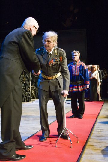 Sotilaspukuinen mies, jolla on mitalit ja joka käyttää keppiä, kättelee toista juhlapukuista miestä punaisella matolla. Taustalla seisoo muita ihmisiä, muun muassa perinteiseen asuun pukeutunut henkilö.