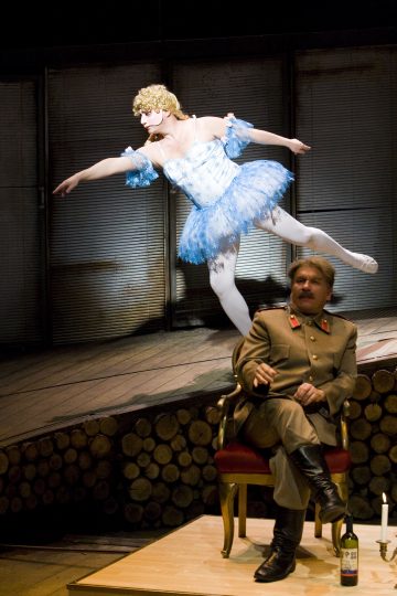 Sotilaspukuinen mies istuu valtaistuimen kaltaisella tuolilla, ja hänen vieressään on viinipullo, kun taas siniseen tutuun ja valkoiseen naamariin pukeutunut ballerina tanssii energisesti taustalla.