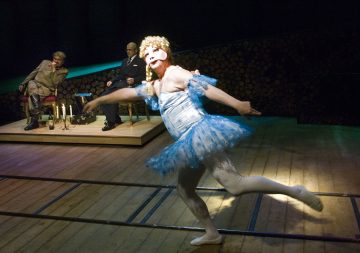 Siniseen tutuun ja valkoisiin sukkahousuihin pukeutunut esiintyjä tanssii puisella lavalla, kun kaksi pukumiestä istuu ja katselee taustalla kynttilöiden valaisemalta lavalta.