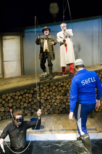 Kaksi vanhempaa miestä takissa ja saappaissa seisoo puisella laiturilla kalastamassa, ja lähellä seisoo mies, jolla on 