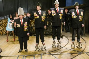 Neljä pukuista, suuriin hattuihin ja moniin mitaleihin pukeutunutta miestä seisoo metallirunkoisilla korokkeilla puulavalla ja nostaa yhtä kättä. Kaksi naista seisoo taustalla pinottujen polttopuiden vieressä. Kohtaus vaikuttaa teatraaliselta ja surrealistiselta.