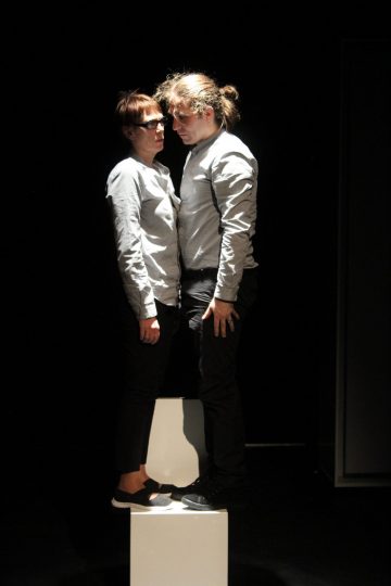 Kaksi ihmistä seisoo tiiviisti vastakkain pienellä valkoisella alustalla pimeässä tilassa, molemmilla on vaaleanharmaat paidat ja tummat housut. He vaikuttavat vakavilta, ja heitä valaisee valokeilan valo.