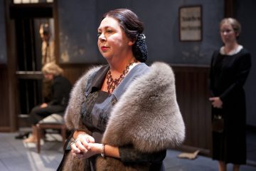 Hämärästi valaistun huoneen etualalla seisoo mietteliään näköinen nainen, jolla on muodollinen mekko ja turkisolki. Taustalla on kaksi naista ja yksi mies, yksi istuu ja kaksi seisoo, kaikki vaikuttavat vakavilta.