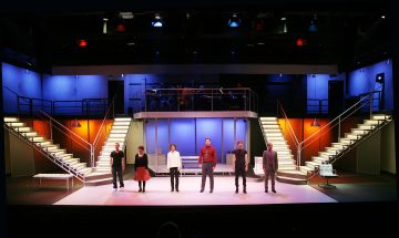 Kuusi ihmistä seisoo erillään toisistaan kirkkaasti valaistulla modernilla teatterilavalla, jossa on värikäs taustavalo, kaksi valkoista portaikkoa molemmin puolin ja korotettu lava, jonka taustalla on soittimia.