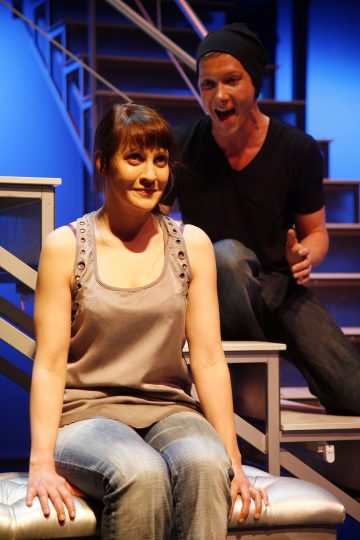 Nainen istuu penkillä hymyillen pehmeästi, kun taas hänen takanaan oleva mustaan paitaan ja pipoon pukeutunut mies näyttää puhuvan energisesti. He ovat lavalla, jonka taustalla on metalliportaat ja sininen valaistus.