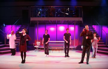 Kuusi näyttelijää seisoo kirkkaasti valaistulla näyttämöllä, jossa on vaaleanpunainen ja violetti valaistus. Takana olevalla korotetulla lavalla soittaa bändi. Näyttelijät ovat kesken esityksen, jotkut elehtivät ja yhdellä on laboratoriotakki.