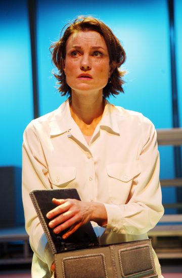 Lyhyet ruskeat hiukset omaava nainen, jolla on valkoinen pusero, katsoo ylöspäin huolestuneena seisoen ja pitäen kädessään avointa laukkua tai salkkua. Sinisävyinen valaistus ja epäselvä tausta viittaavat näyttämöasetelmaan.