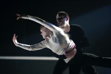 Mies ja nainen esittävät nykytanssia lavalla. Valkoiseen pukeutunut nainen ojentaa käsivartensa sirosti, kun mustiin pukeutunut mies tukee häntä takaapäin. Valaistus on dramaattinen ja korostaa heidän ilmeikkäitä liikkeitään.