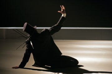 Tummaan pukuun ja silinterihattuun pukeutunut henkilö istuu lattialla dramaattisessa valaistuksessa ja kurottautuu ylöspäin toinen käsi ojennettuna ja sormet levällään, mikä luo teatraalisen ja ilmeikkään asennon.