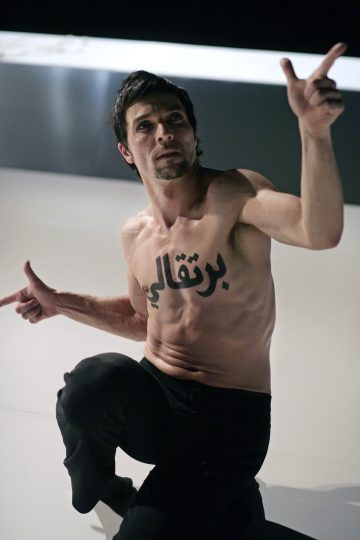 Paidaton mies, jolla on lyhyet tummat hiukset ja suuri arabialainen tatuointi rinnassaan, polvistuu lattialle, osoittaa molemmilla käsillään ja katsoo vakavalla ilmeellä eteenpäin.