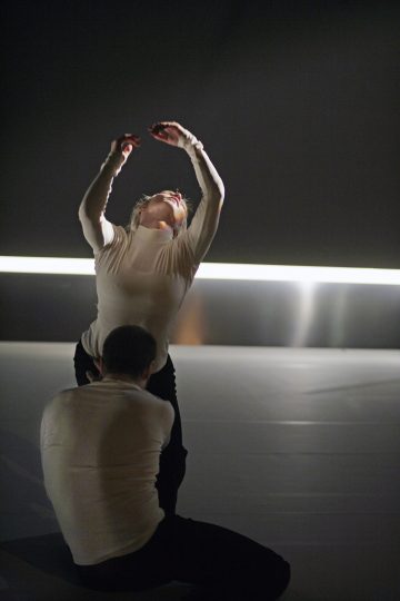 Valkoiseen toppiin pukeutunut henkilö poseeraa kädet kohotettuna ja pää taaksepäin kallistettuna, kun taas toinen, niin ikään valkoiseen toppiin pukeutunut henkilö polvistuu heidän edessään hämärässä taustassa, jossa on vaakasuora valonauha.