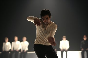 Valkoiseen pitkähihaiseen paitaan ja mustiin housuihin pukeutunut henkilö esittää nykytanssiliikkeen dramaattisessa valaistuksessa, ja taustalla istuu useita ihmisiä.