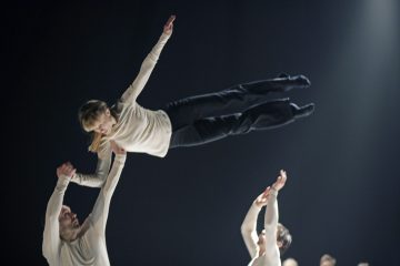 Ryhmä tanssijoita esiintyy lavalla. Yksi tanssija on keskellä ilmaa, jota toinen tanssija pitää vaakasuorassa toisessa ojennetussa kädessä, ja muut tanssijat kurottelevat ylöspäin. Lava on hämärästi valaistu, mikä korostaa heidän koordinoitua liikettään.