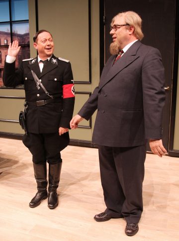 Kaksi näyttelijää näyttämöllä; toisella on musta natsien SS-puku ja hän nostaa kätensä ylös, kun taas toinen seisoo vieressä isossa harmaassa puvussa, jolla on silmälasit ja parta. He näyttävät esiintyvän teatterinäytelmässä.