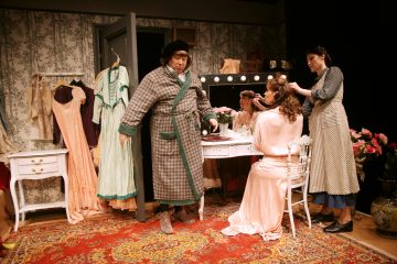 Kolme ihmistä on vintage-tyylisessä pukuhuoneessa. Yksi nainen istuu turhamaisuuden ääressä, kun toinen muotoilee hiuksiaan. Lähistöllä seisoo mies, jolla on aamutakki ja hattu. Huone on koristeltu mekoilla, kukilla ja koristeellisella matolla.