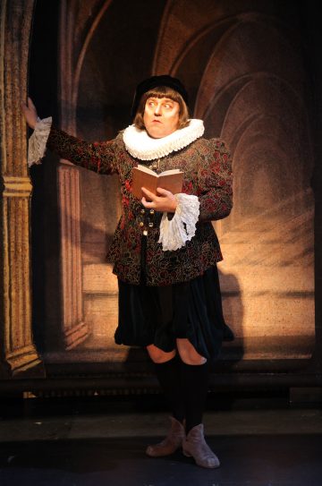 Historialliseen renessanssipukuun pukeutunut henkilö, jolla on röyhelöinen kaulus, tumma hattu ja kirjailtu takki, seisoo kaaripylvään vieressä pitäen kirjaa kädessään ja ilmeikäs ilme kasvoillaan.