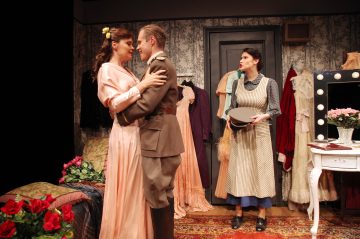 Vaaleanpunaiseen mekkoon pukeutunut nainen syleilee sotilasunivormuun pukeutunutta miestä, kun toinen esiliinaan pukeutunut nainen seisoo vieressä hattu kädessään, vintage-tyylisessä huoneessa, jossa on kukkia, mekkoja ja valaistu turhamaisuuspeili.