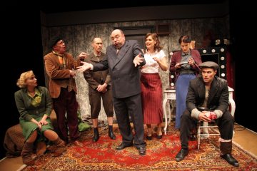 Seitsemän vintage-asuihin pukeutunutta näyttelijää esiintyy lavalla huoneessa, jossa on kukkatapetti ja koristeellinen matto; yksi pukumies elehtii dramaattisesti, kun muut reagoivat erilaisin ilmein ja asennoin.