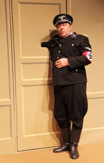 Mustaan natsiunivormuun ja lippalakkiin pukeutunut mies seisoo ruskehtavaa ovea vasten, hänen vasen kätensä on taivutettu seinän taakse aukon läpi, ja hän näyttää yllättyneeltä tai huolestuneelta. Hänellä on punainen käsivarsinauha, jossa on hakaristi.