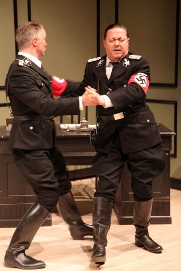 Kaksi natsitason SS-univormuun pukeutunutta miestä, joilla on hakaristipäivän käsivarsinauha, esittää teatterikohtauksen, jossa he pitävät toisiaan kädestä kiinni ja näyttävät tanssivan tai kamppailevan toimistomaisessa ympäristössä olevan työpöydän edessä.