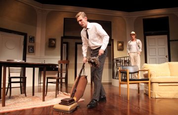 Valkoiseen paitaan ja solmioon pukeutunut mies imuroi lattiaa vintage-tyylisessä olohuoneessa, kun taas toinen henkilö, jolla on rennot vaatteet ja hattu, seisoo taustalla oven lähellä.