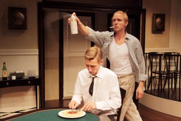 Valkoiseen paitaan ja solmioon pukeutunut mies syö spagettia pöydässä, kun toinen mies, jolla on rennot vaatteet ja savuke suussaan, suihkuttaa jotain ruoan päälle seisoen hänen takanaan dramaattisessa kohtauksessa.