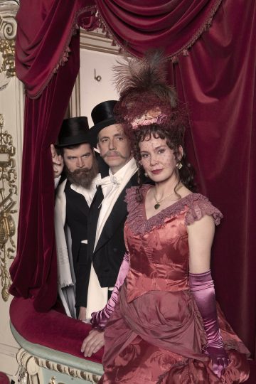 Kolme aikakauden pukuihin pukeutunutta ihmistä seisoo ylellisessä, punasamettisessa teatterilokerossa. Kaksi mustiin pukuihin ja silinterihattuihin pukeutunutta miestä katselee ulos edessä itsevarmasti seisovan, punaiseen viktoriaaniseen mekkoon ja hienostuneeseen hattuun pukeutuneen naisen takaa.