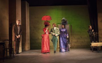 Viisi näyttelijää lavalla aikakauden puvuissa: mies seisoo yksin vasemmalla, kirkkaanpunaiseen mekkoon ja hattuun pukeutunut nainen pitelee punaista sateenvarjoa, kaksi muuta vihreään ja violettiin pukeutunutta kävelee yhdessä, ja toinen seisoo verhon lähellä oikealla.