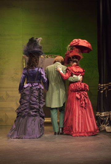 Kolme hienostuneisiin aikakauden pukuihin pukeutunutta ihmistä seisoo takaapäin katsottuna käsi kädessä lavalla. Kahdella on pitkät mekot ja hattu - toisella violetti, toisella punainen - ja keskimmäisellä henkilöllä on vihreä ruudullinen puku.