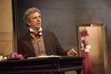 Aikakauden vaatteisiin pukeutunut mies istuu pöydän ääressä ja katsoo sivulle. Pöydällä on violetteja sipuleita ja papereita. Taustalla punamekkoon ja hattuun pukeutunut nainen seisoo peilin edessä hieman epätarkasti.