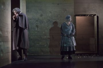 Tummaan viittaan pukeutunut mies seisoo seinän vieressä mietteliään näköisenä, kun taas vanhanaikaisiin vaatteisiin ja huiviin pukeutunut nainen seisoo veitsi kädessään tyhjän kuvakehyksen vieressä hämärästi valaistulla näyttämöllä.