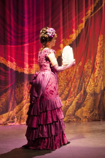 Nainen, jolla on koristeellinen magentanpunainen viktoriaanisen tyylin mekko, seisoo lavalla valkoista viuhkaa kädessään, ja taustalla on dramaattinen punakultainen verho.
