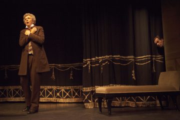 Aikakauden pukuihin pukeutunut mies seisoo lavalla rintaansa puristellen ja ahdistuneen näköisenä, kun taas toinen mies kurkistaa ulos raskaan verhon takaa. Näyttämö on hämärästi valaistu, ja siellä on penkki ja koristeellinen tumma tausta.