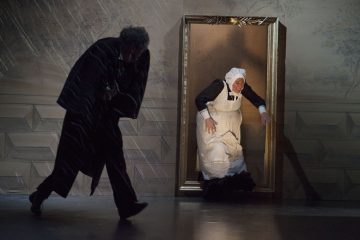 Kaksi näyttelijää esiintyy näyttämöllä; toinen on pukeutunut hattuun ja esiliinaan pukeutuneeksi piiaksi, joka astuu ulos ovesta tai kuvakehyksestä, kun taas toinen tummiin vaatteisiin pukeutunut näyttelijä kumartuu ja kävelee pois dramaattisessa valaistuksessa.