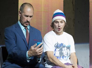 Siniseen pukuun pukeutunut mies näyttää puhelintaan nuoremmalle miehelle, jolla on valkoinen graafinen T-paita ja raidallinen pipo; molemmat näyttävät yllättyneiltä ja keskittyvät näyttöön.