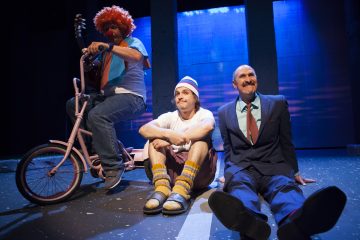 Lavalla istuu kolme pukumiestä sinisessä valaistuksessa. Yhdellä on punainen peruukki ja hän istuu vaaleanpunaisella kolmipyöräisellä polkupyörällä, toinen pipo päässä ja epäsopivissa vaatteissa istuu ristissä ja kolmas pukumies hymyilee lattialla istuen.