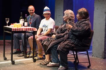 Neljä ihmistä istuu pöydän ympärillä, ja lavalla on viiniä ja juustoa. Kahdella on raidallinen kaapu, yhdellä puku, ja yksi on pukeutunut rennosti hattuun ja värikkäisiin sukkiin. He näyttävät keskustelevan teatteriesityksen aikana.