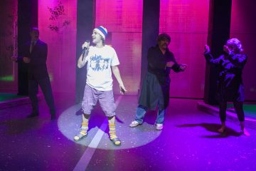 Neljä ihmistä esiintyy lavalla, jossa on värikäs ja dramaattinen valaistus. Edessä olevalla henkilöllä on pipo, shortsit ja bändin t-paita päällään, ja hän laulaa mikrofoniin; kolme muuta on taustalla, ja he näyttelevät erilaisissa asennoissa.